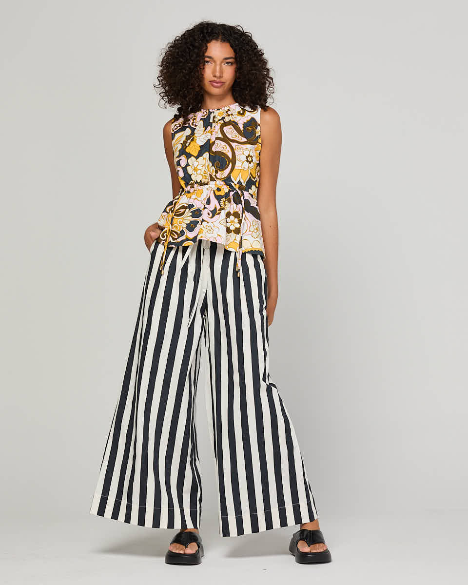 A model wearing a sleeveless floral top with yellow, black, and white patterns, paired with black and white vertically striped wide-leg pants and black platform sandals, set against a plain light gray studio background.