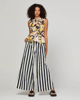 A model wearing a sleeveless floral top with yellow, black, and white patterns, paired with black and white vertically striped wide-leg pants and black platform sandals, set against a plain light gray studio background.
