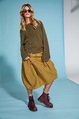 Woman wearing a khaki jumper and mustard plaid skirt with maroon boots on a blue background