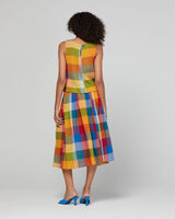 A woman stands with her back to the camera wearing a sleeveless top and skirt with a colorful striped bodice and a multicolor plaid skirt, paired with blue heels, against a light gray studio background.
