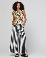 A model wearing a sleeveless top with a black, gold, and white paisley pattern paired with black and white vertically striped wide-leg pants, standing against a light gray studio background.