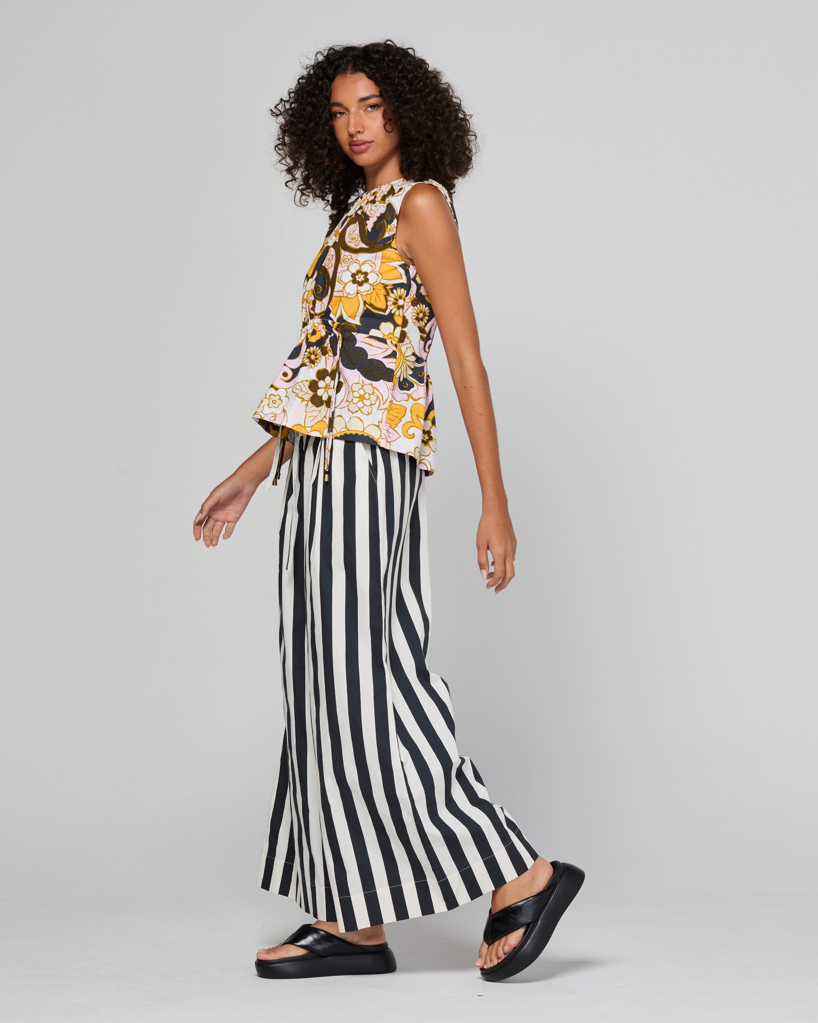 A model wearing a sleeveless floral top with yellow, black, and white patterns, paired with black and white vertically striped wide-leg pants and black platform sandals, set against a plain light gray studio background.