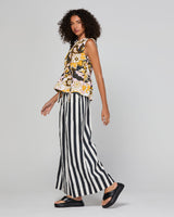 A model wearing a sleeveless floral top with yellow, black, and white patterns, paired with black and white vertically striped wide-leg pants and black platform sandals, set against a plain light gray studio background.