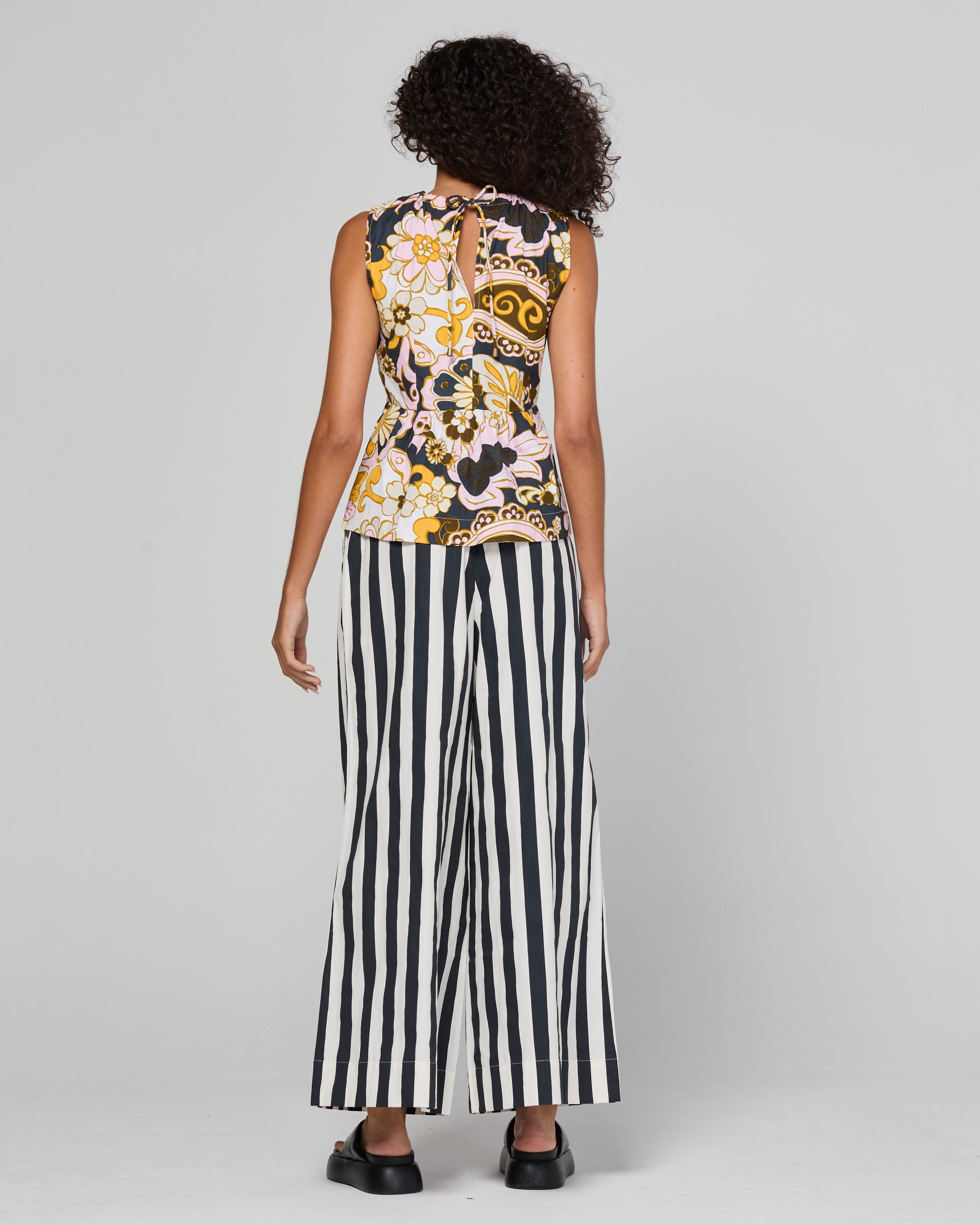 A model shown from the back wearing a sleeveless top with a colorful floral and abstract print and black-and-white vertically striped wide-leg pants, against a light gray studio background.