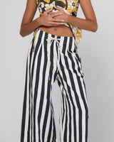 A model wearing high-waisted, wide-leg pants with bold vertical black and white stripes, paired with a patterned crop top, against a neutral gray background.