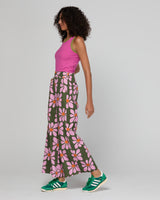 Woman wearing a pink top and floral pants with green sneakers on a plain background