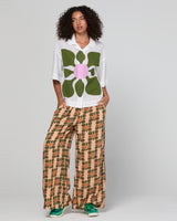 Woman wearing a white shirt with a large green floral design and patterned pants on a plain background