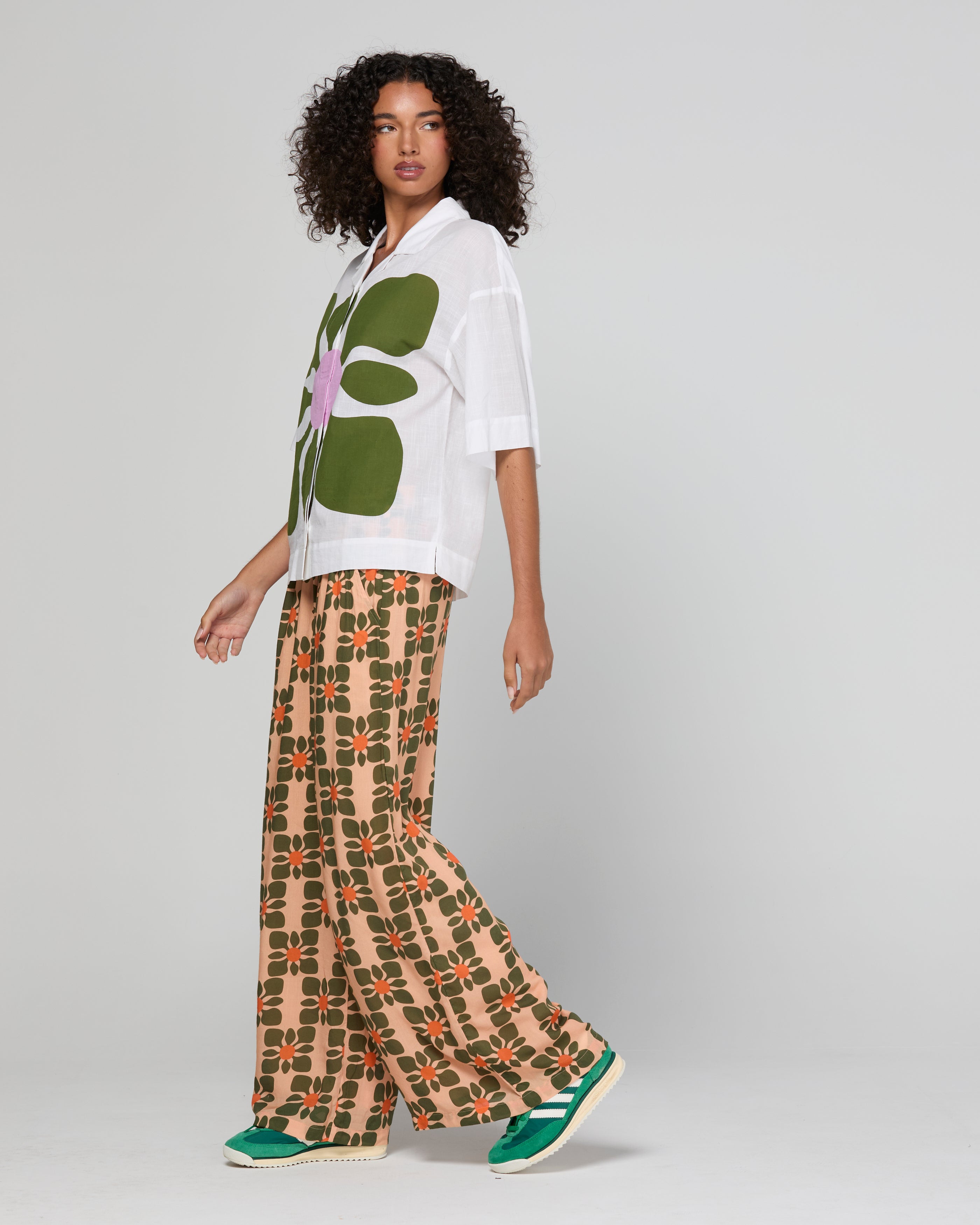 Woman wearing a white shirt with a green floral design and patterned pants on a gray background