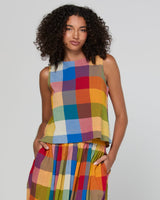 A model with curly dark hair wearing a sleeveless top and matching pleated skirt set in a vibrant multicolor plaid pattern, set against a plain light gray studio background.