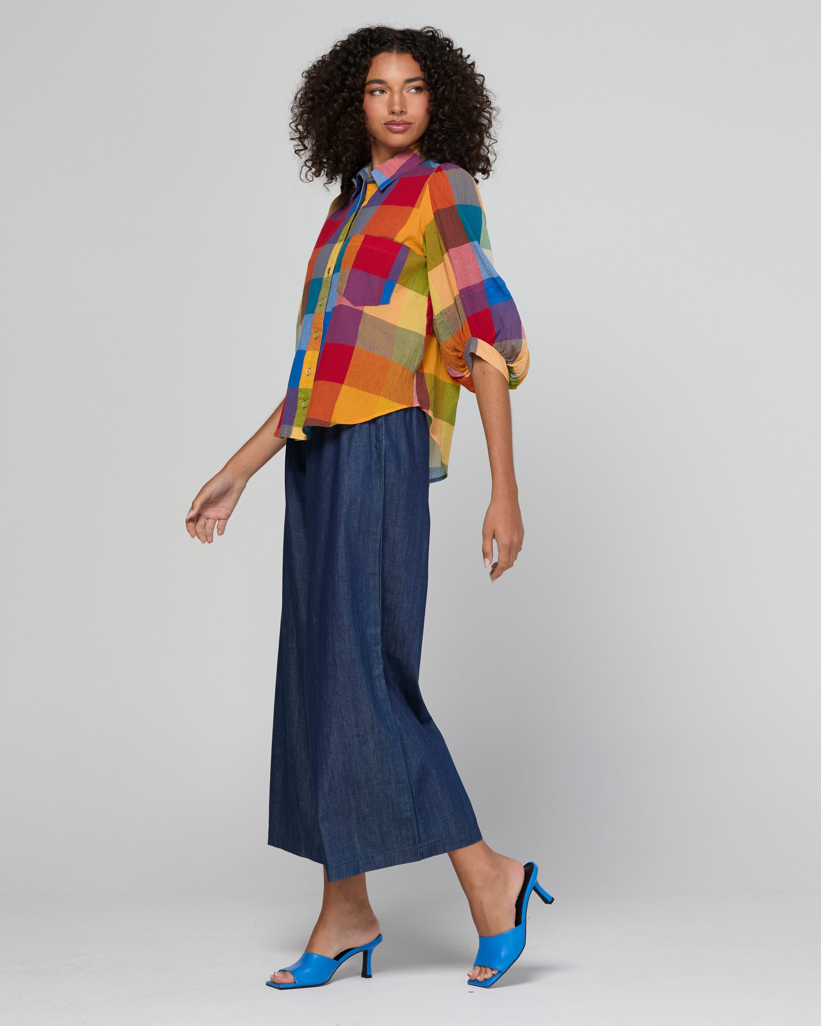 A model is shown from the side wearing a vibrant multicolor plaid shirt with three-quarter sleeves and dark blue wide-leg cropped pants, paired with bright blue heeled sandals, against a light gray studio background.