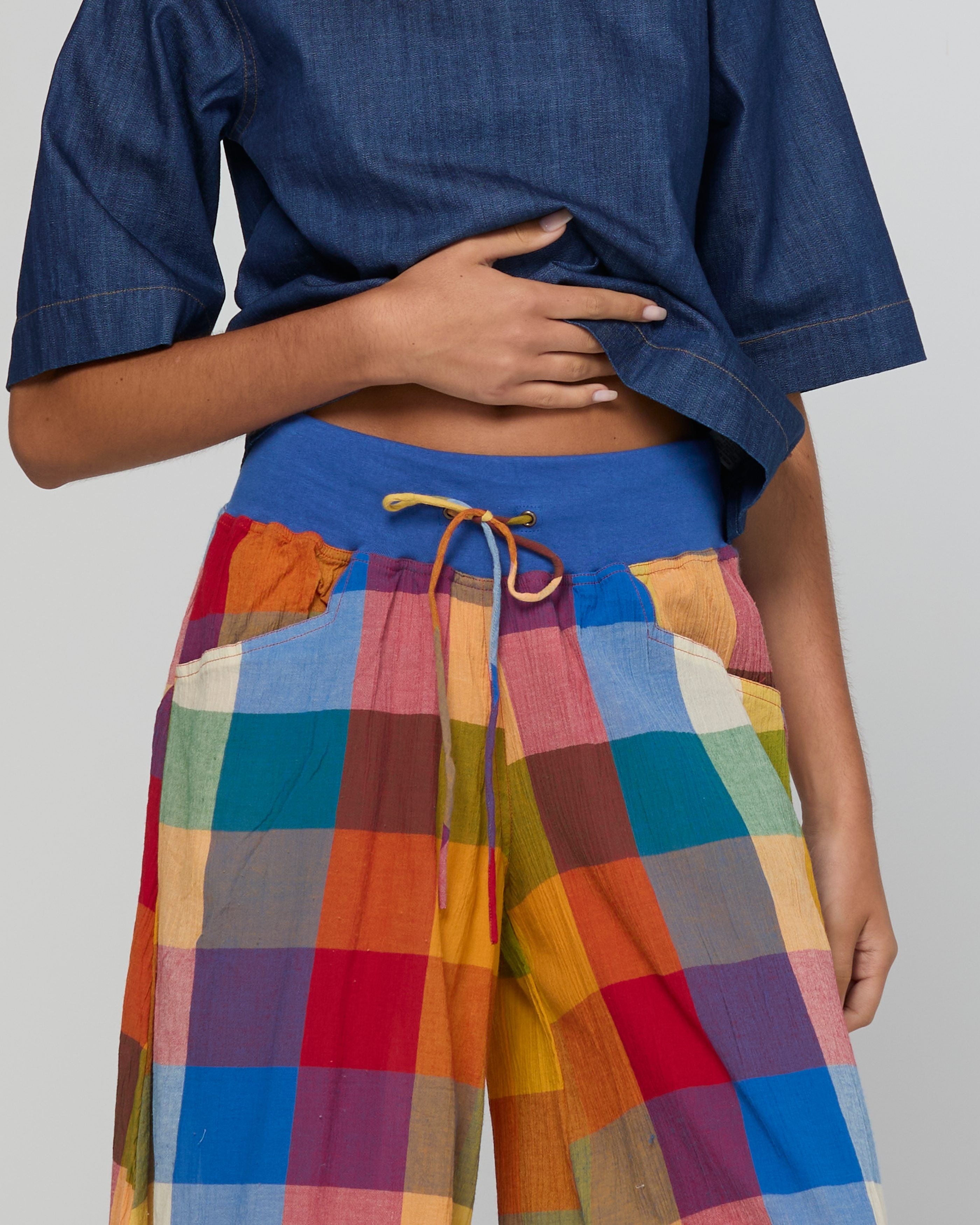A close-up of a person wearing colorful patchwork plaid pants with a blue waistband and drawstring, paired with a blue short-sleeve top against a light gray studio background.