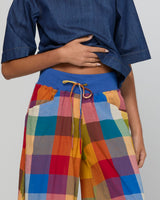 A close-up of a person wearing colorful patchwork plaid pants with a blue waistband and drawstring, paired with a blue short-sleeve top against a light gray studio background.
