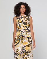 A woman with curly dark hair models a sleeveless jumpsuit featuring a bold, retro paisley and floral pattern in yellow, brown, black, and white against a light gray studio background.