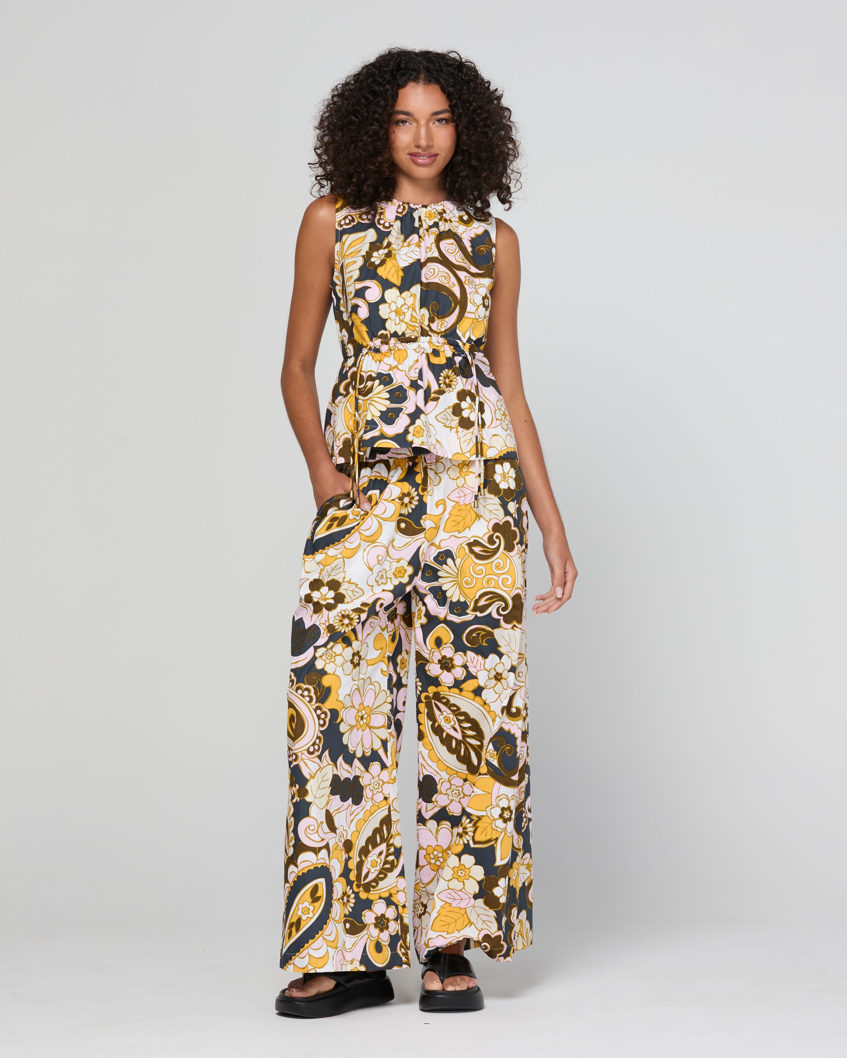 A model wearing a sleeveless top with a wide-leg pant and a peplum waist, featuring a yellow, black, and white paisley floral pattern, photographed against a light gray studio background.