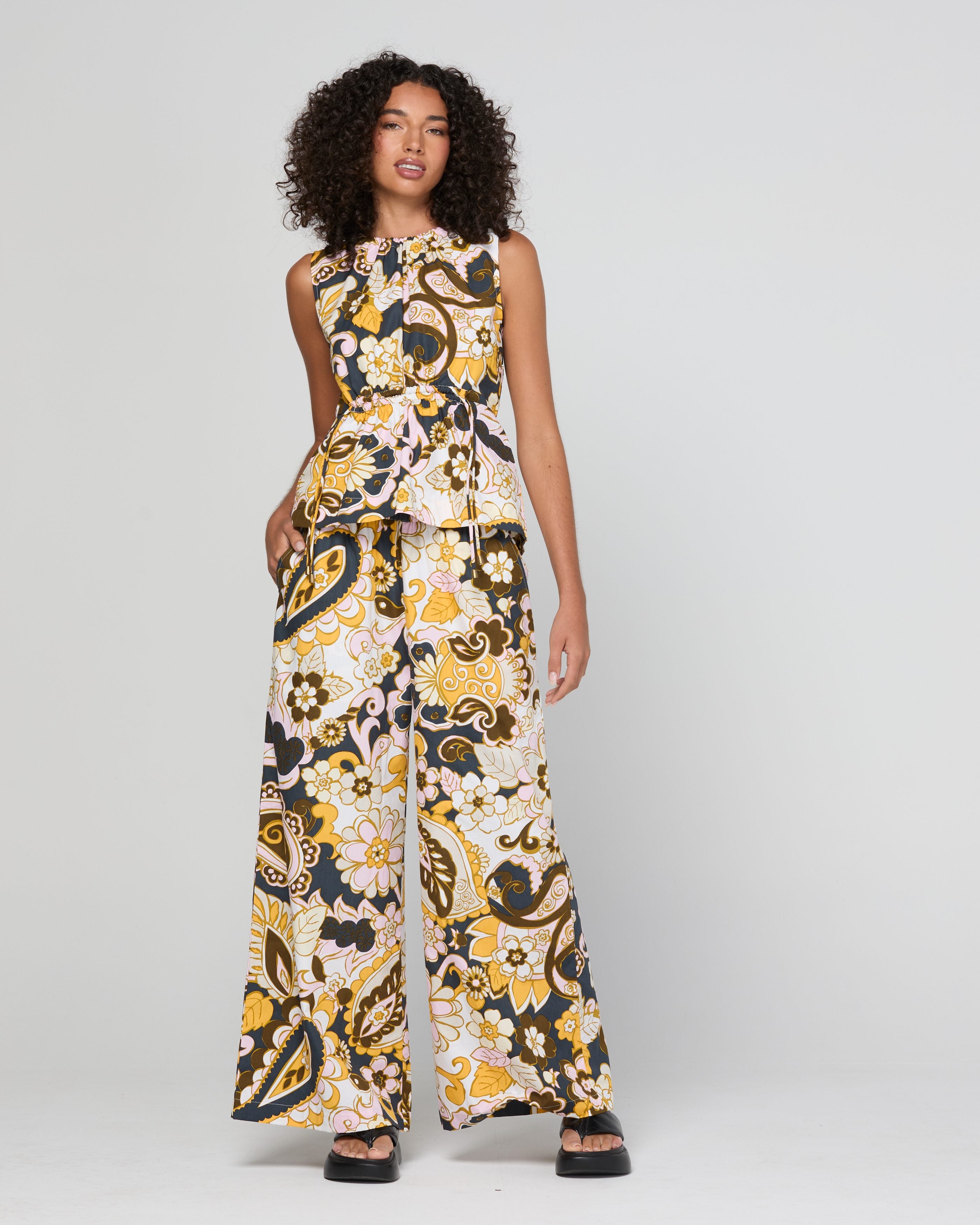 A woman modeling a sleeveless, wide-leg jumpsuit with a bold paisley pattern in black, yellow, and cream, set against a plain light gray studio background.