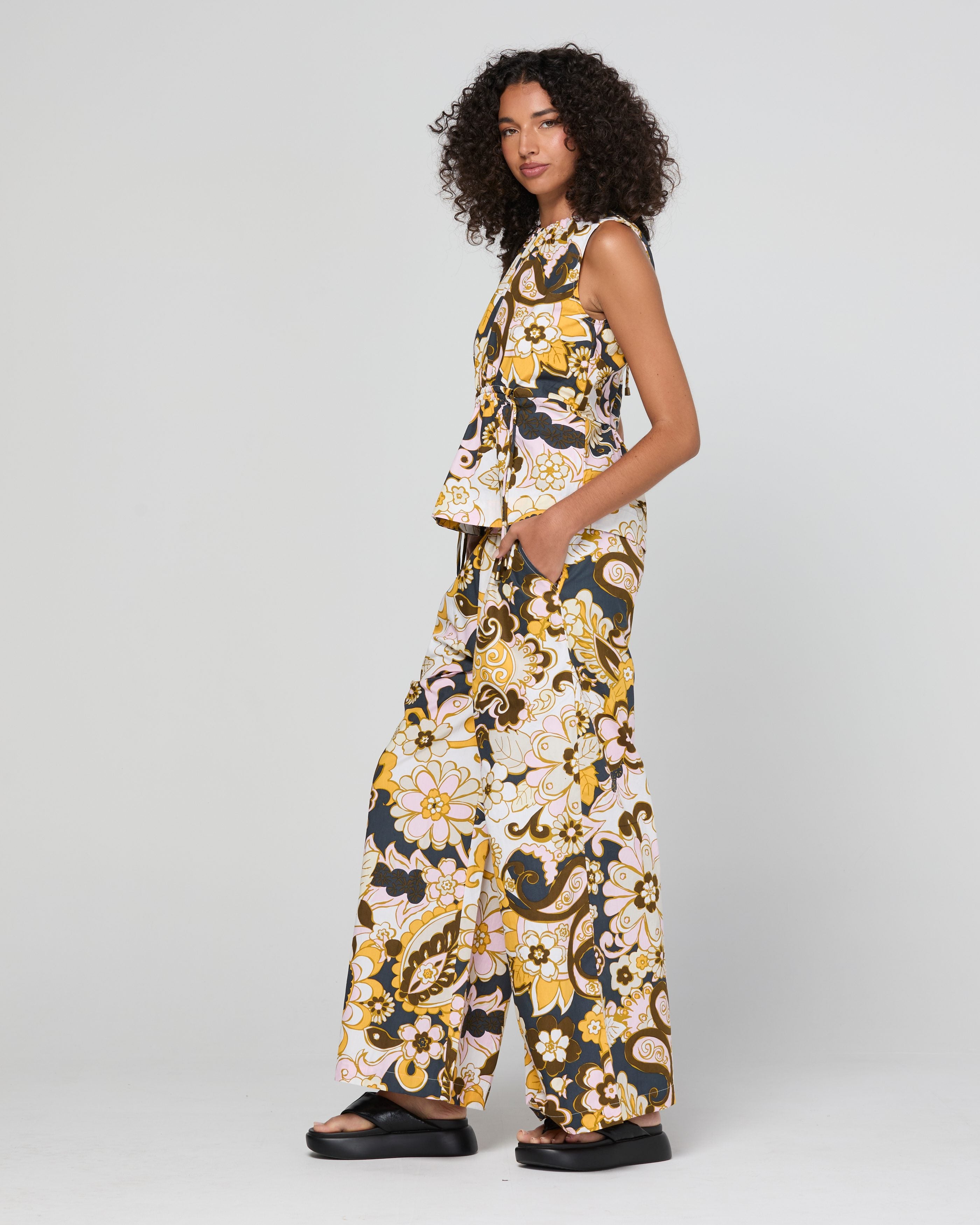 A model wearing a sleeveless matching set with a wide-leg pant and a ruffled peplum top, featuring a bold yellow, black, and white floral pattern, photographed against a light gray studio background.