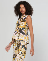 A model with curly dark hair is shown from the side, wearing a sleeveless matching two-piece set featuring a high-neck top and trousers with a bold floral and abstract pattern in yellow, brown, black, and white.