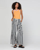 A woman modeling a sleeveless tank top in mandarin colour with light pink trim, paired with black and white vertically striped wide-leg pants, against a light gray studio background.
