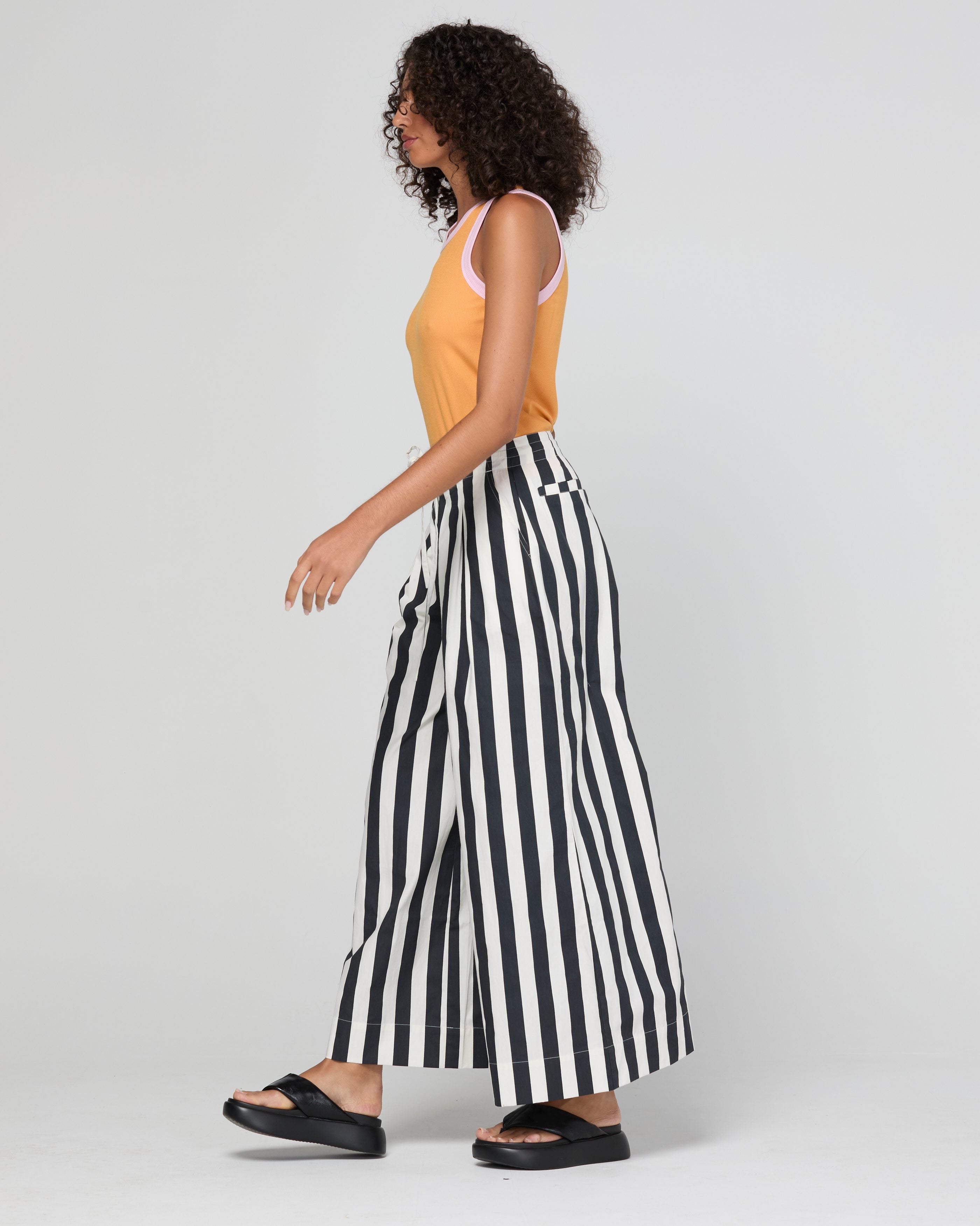 A woman modeling a sleeveless tank top in mandarin colour with light pink trim, paired with black and white vertically striped wide-leg pants, against a light gray studio background.