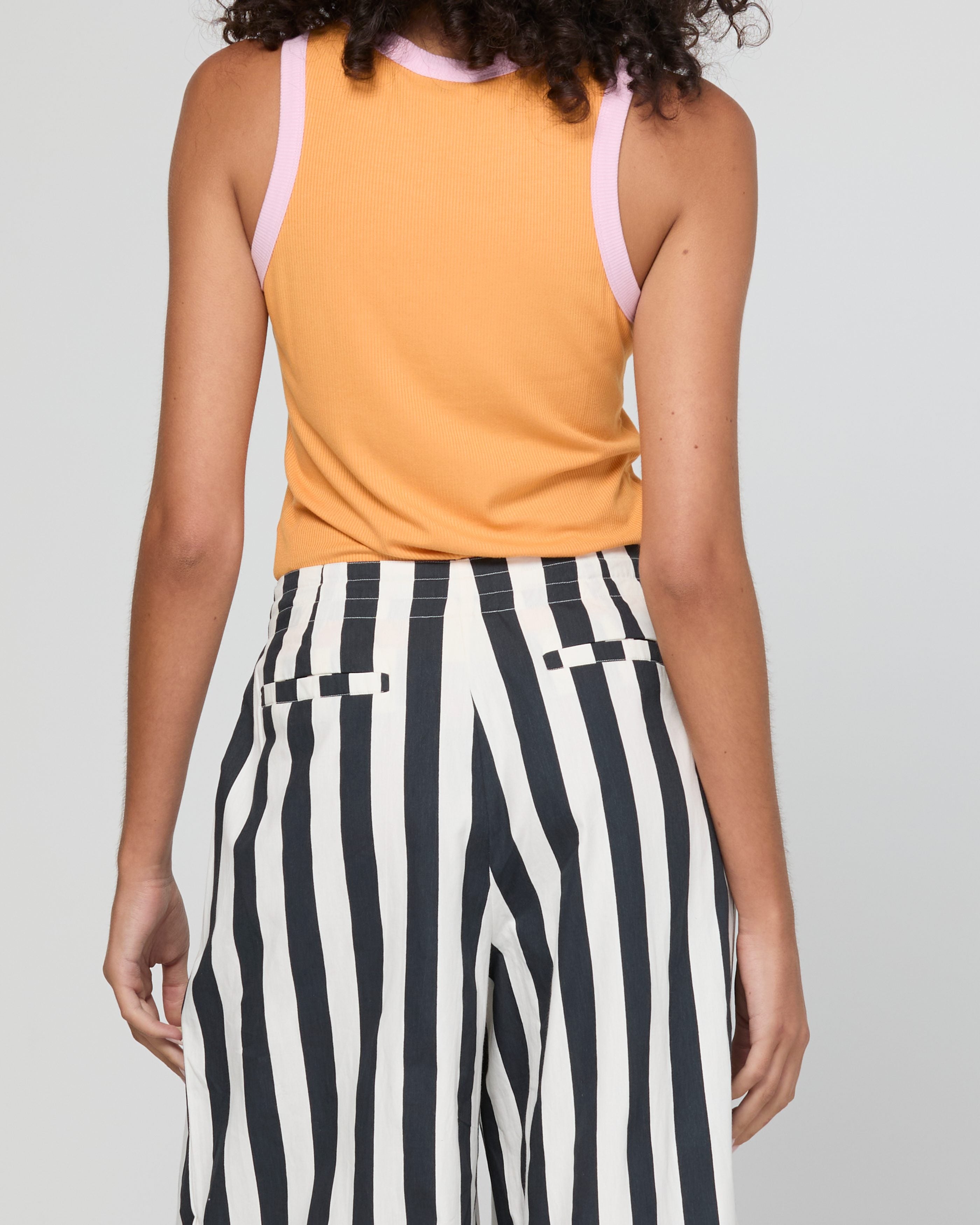 A person shown from the back wearing a sleeveless orange top with light pink shoulder straps and black-and-white vertically striped pants against a light gray studio background.