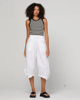 A model stands against a light gray studio background wearing a black and white horizontally striped sleeveless top, loose white cropped pants, and black slide sandals.