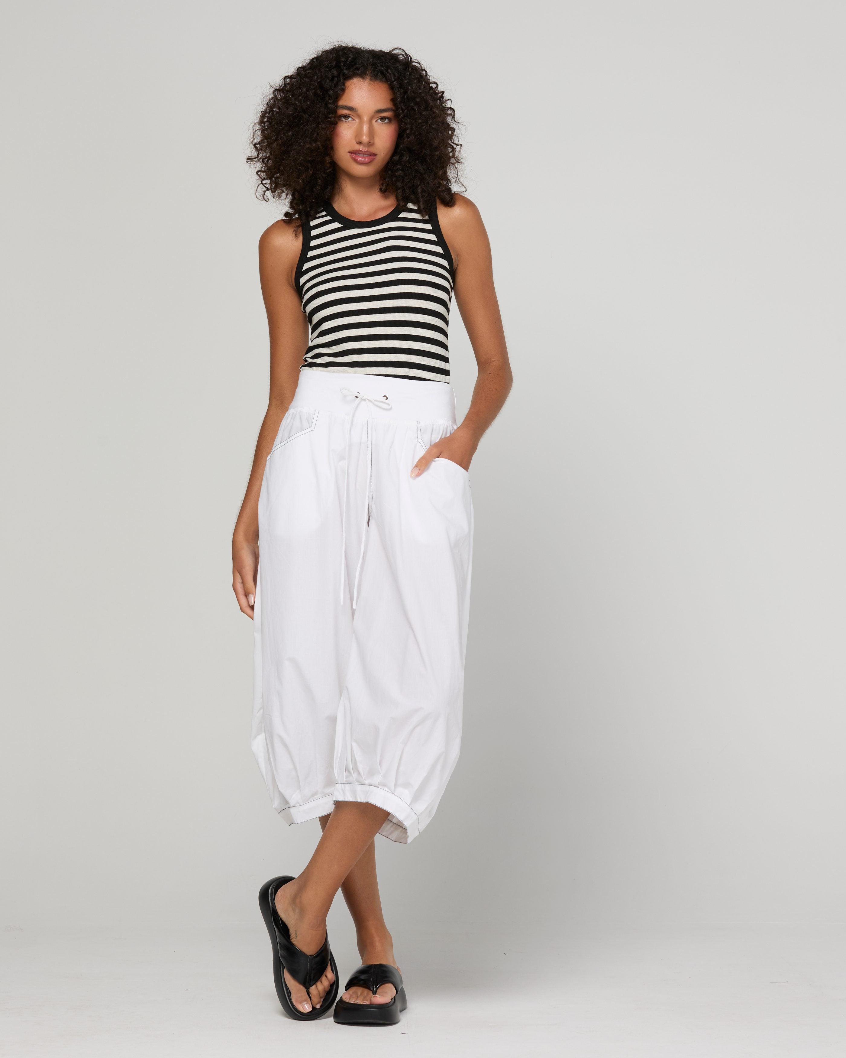Woman wearing a black and white striped tank top and white pants on a gray background
