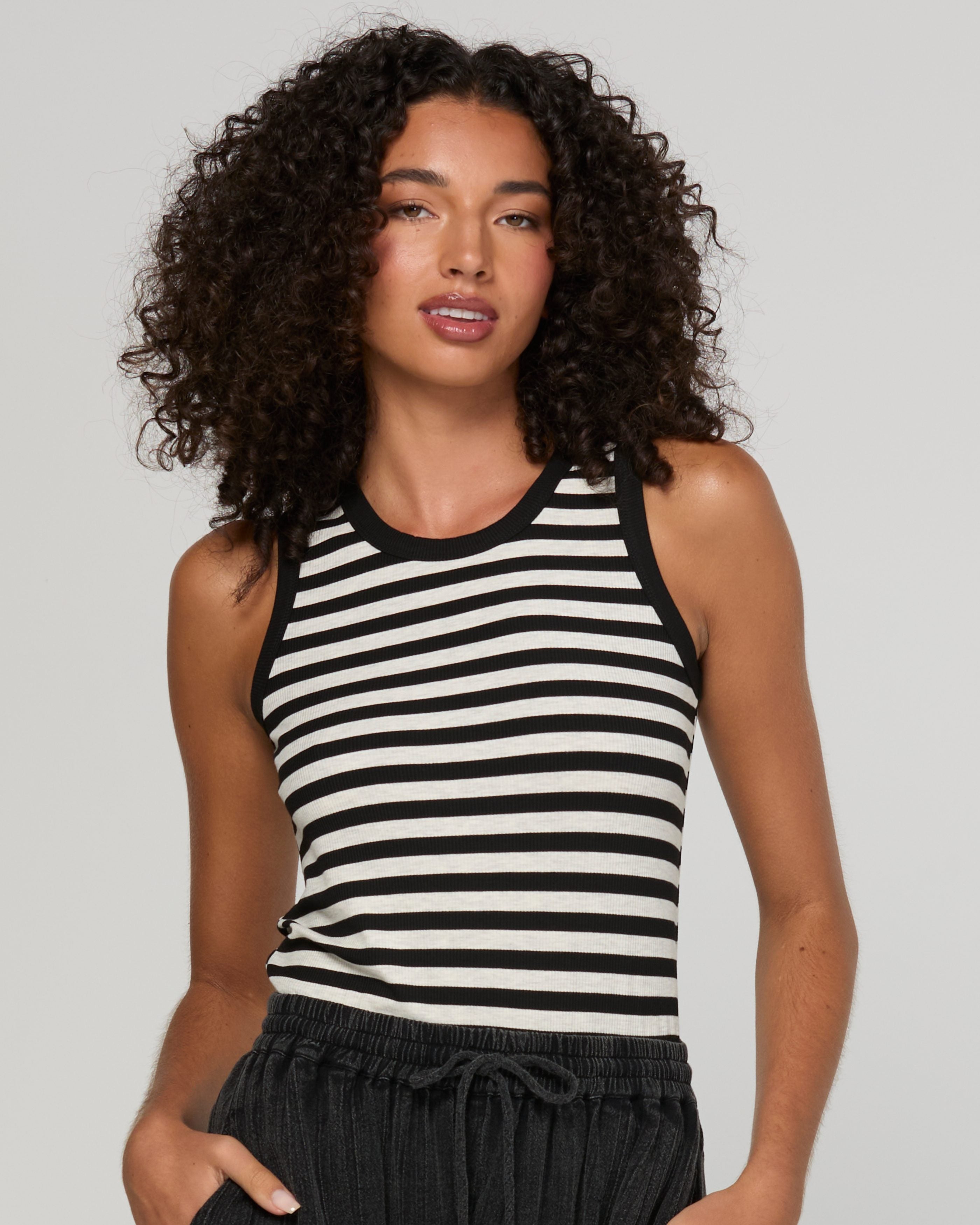 A woman with curly dark hair wearing a black and white horizontally striped ribbed tank top and dark drawstring pants, photographed against a light gray studio background.