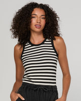 A woman with curly dark hair wearing a black and white horizontally striped ribbed tank top and dark drawstring pants, photographed against a light gray studio background.