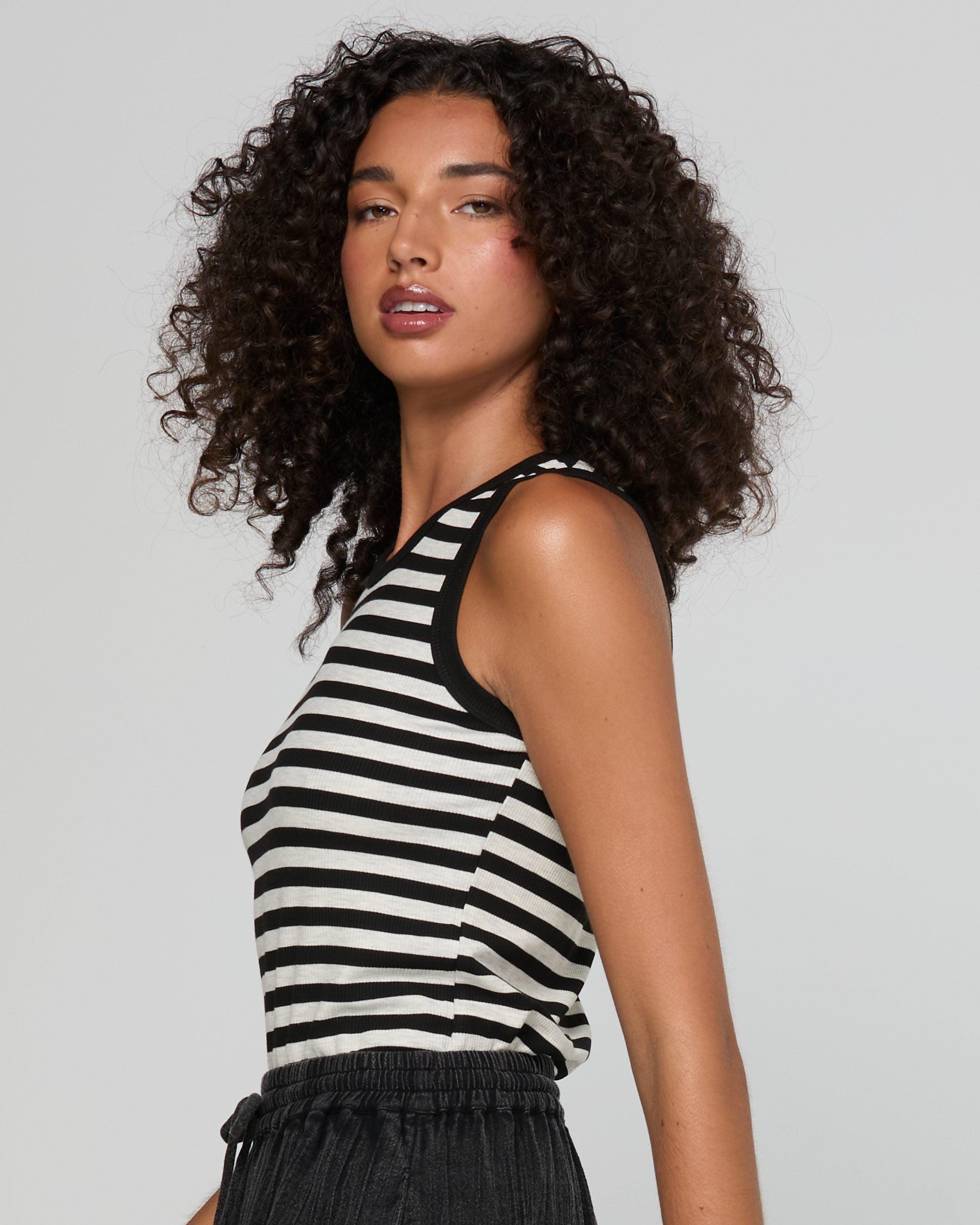 A woman with curly dark hair wearing a black and white horizontally striped sleeveless tank top, shown from a side-back angle against a light gray studio background.