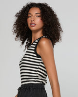 A woman with curly dark hair wearing a black and white horizontally striped sleeveless tank top, shown from a side-back angle against a light gray studio background.