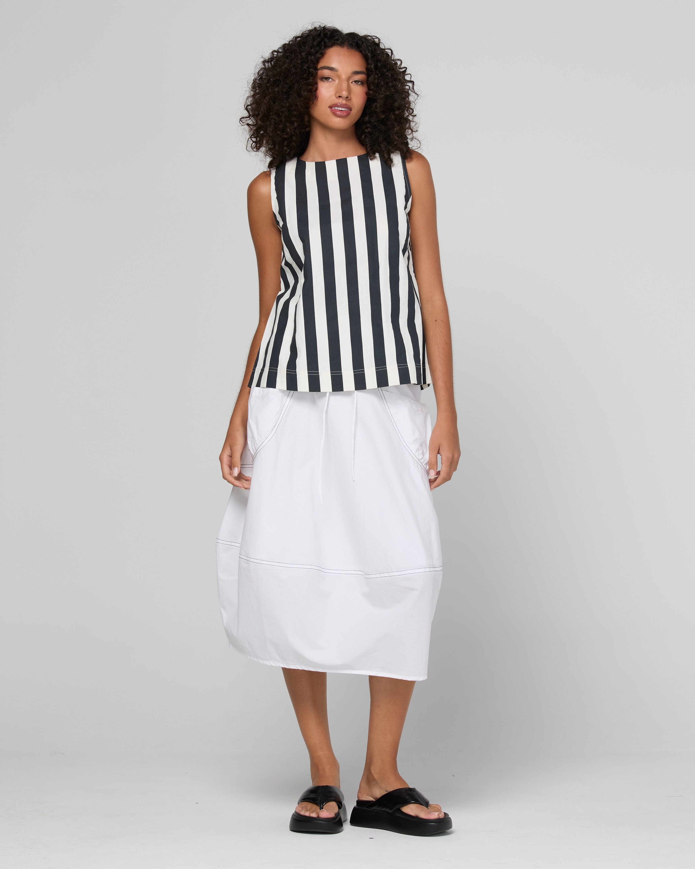 A model wearing a sleeveless top with black and white vertical stripes paired with a white midi skirt and black slide sandals, photographed against a light gray studio background.