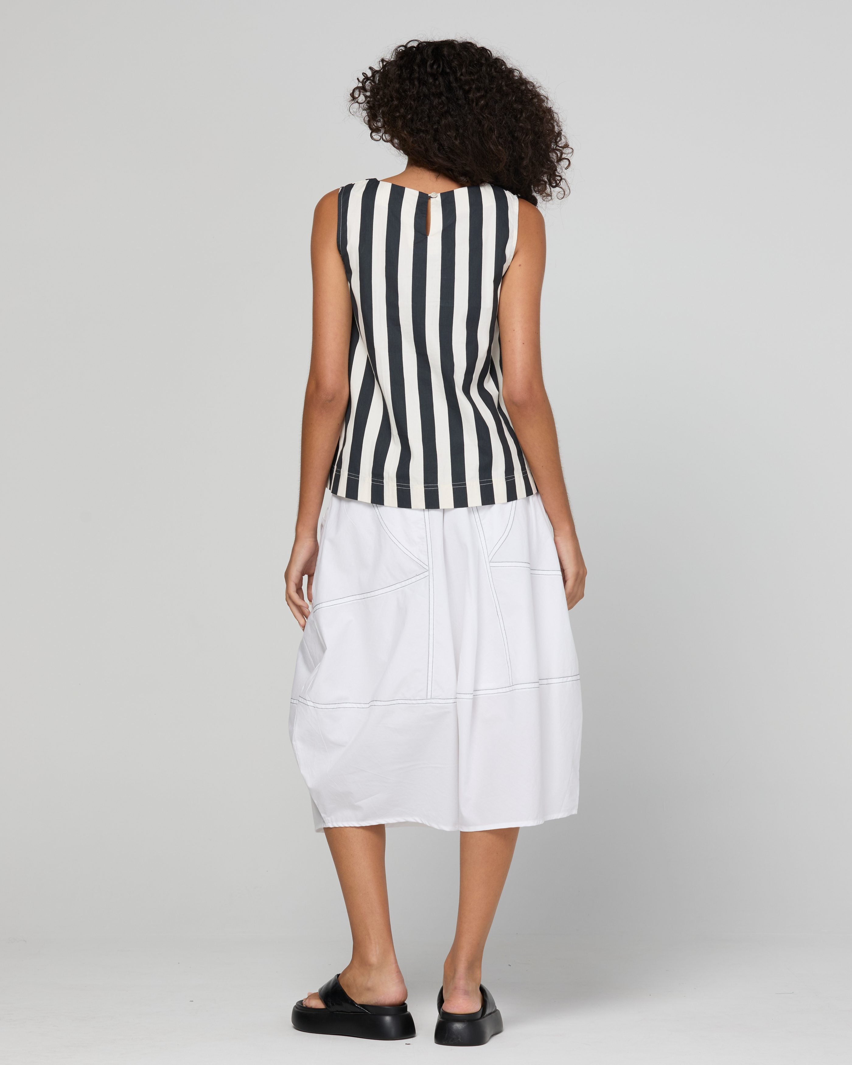 A woman shown from the back wearing a sleeveless black and white vertically striped top paired with a white midi skirt and black platform sandals, against a light gray studio background.