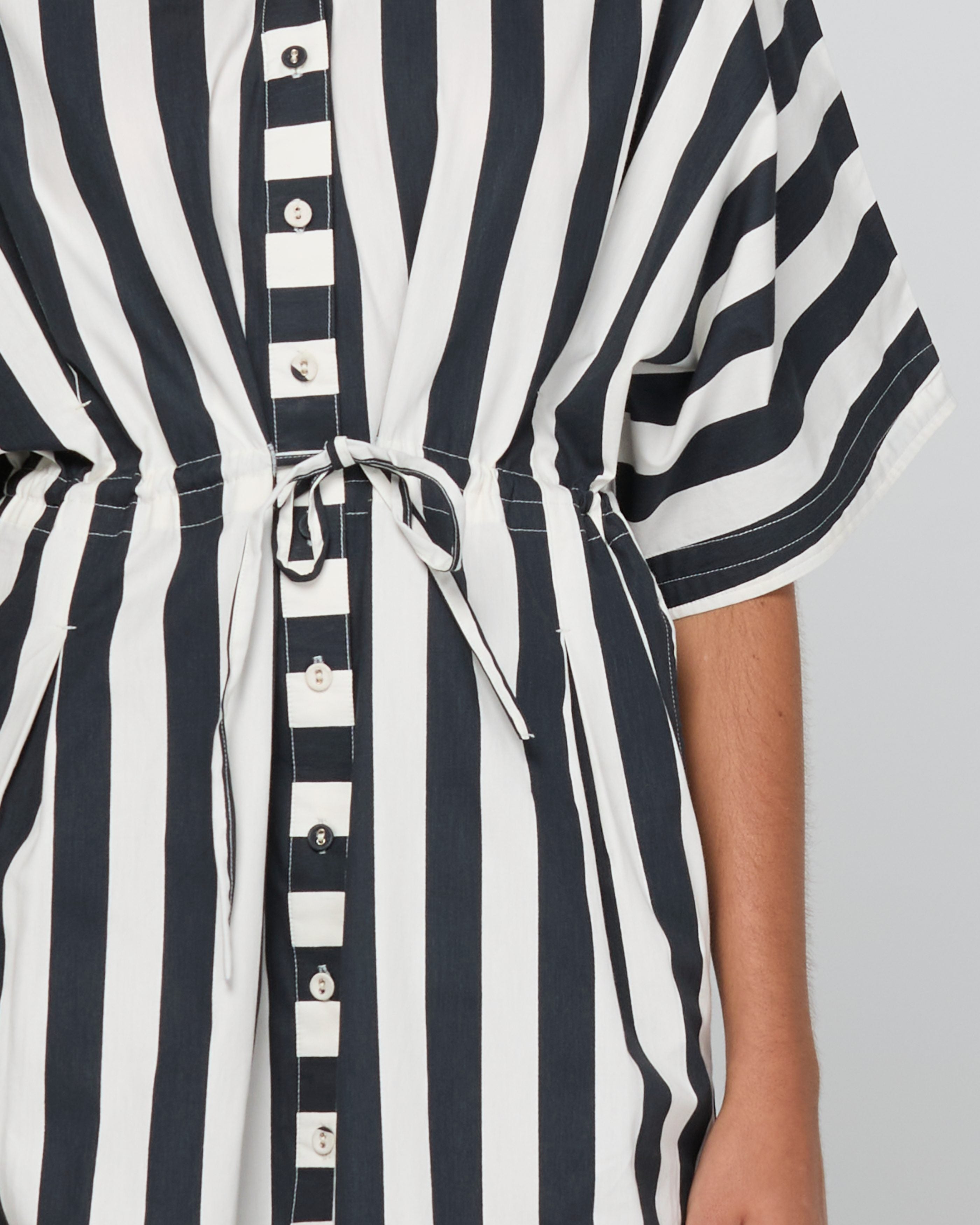 Close-up of a black and white vertically striped short-sleeve shirt dress with a drawstring waist and button front, worn by a model.
