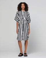 A model wearing a black and white vertically striped shirt dress with a button-down front and a matching tie belt at the waist, paired with black slide sandals, against a light gray studio background.