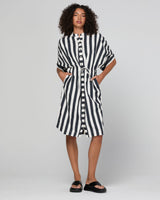 A model wearing a black and white vertically striped short-sleeve button-up shirt dress with a tie waist and side pockets, paired with black slide sandals, against a light gray studio background.