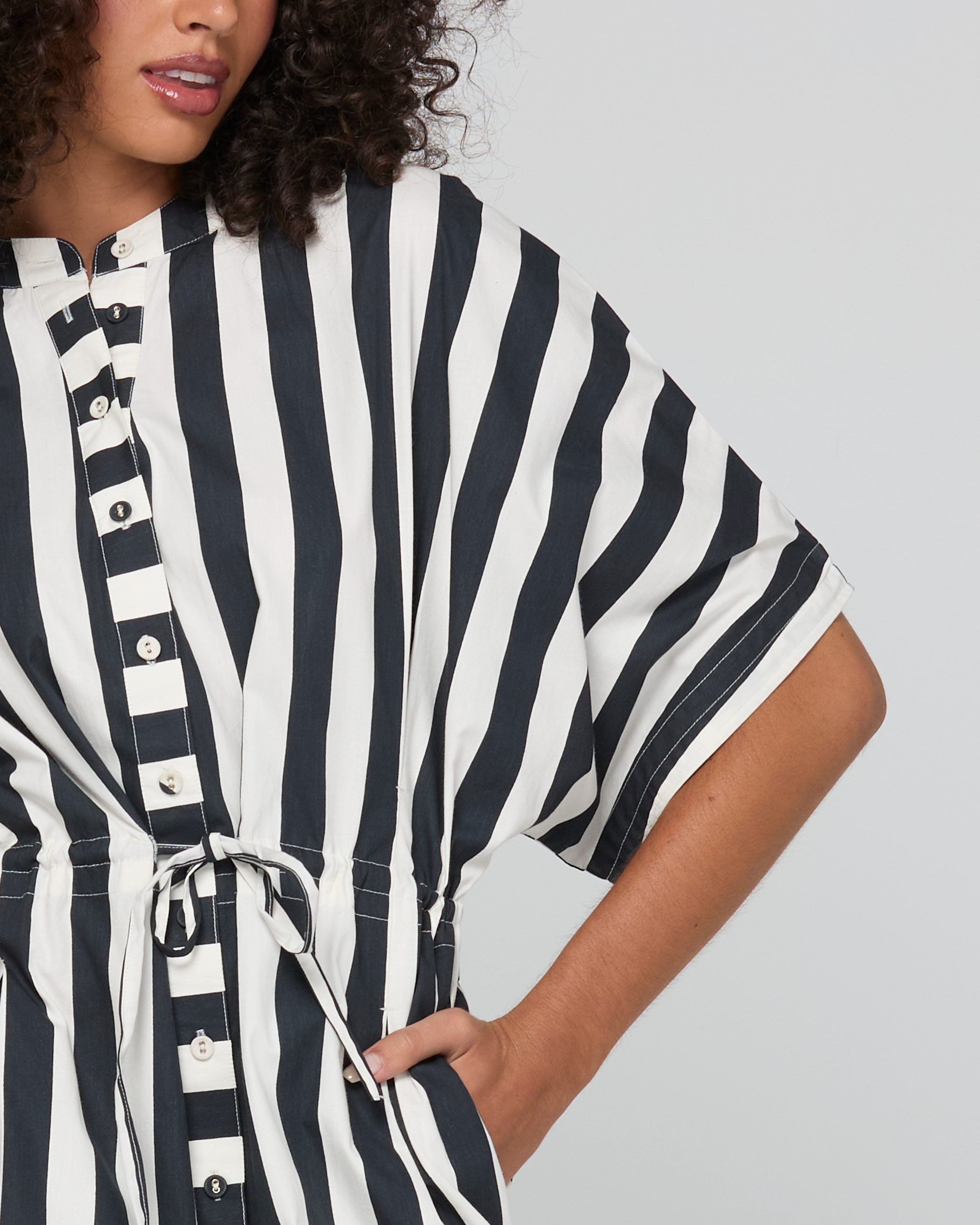 A close-up of a woman wearing a black and white vertically striped short-sleeve tunic top with a buttoned front and tie waist, against a light gray background.