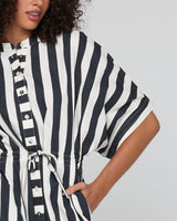 A close-up of a woman wearing a black and white vertically striped short-sleeve tunic top with a buttoned front and tie waist, against a light gray background.