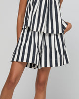 A close-up of a model wearing a matching black and white vertically striped top and shorts set, showing the layered peplum-style top and flared shorts against a plain light gray background.