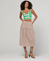 A model stands against a light gray studio background wearing a green and white polka-dot crop top with beige and white gingham tiered midi skirt and green strappy high-heeled sandals.