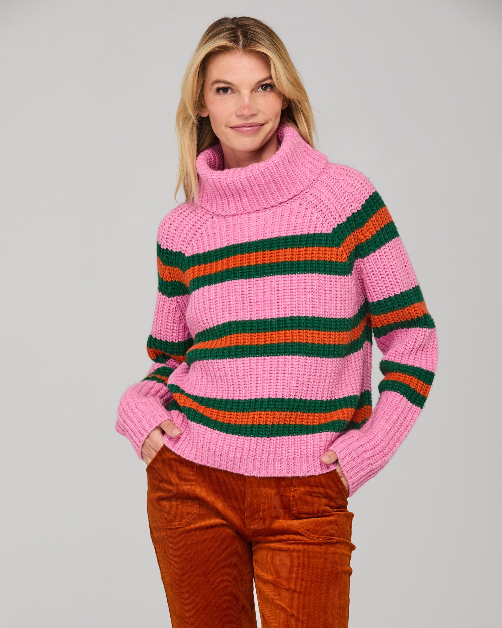 Woman wearing a chunky knit pink jumper with green and orange stripes on a gray background