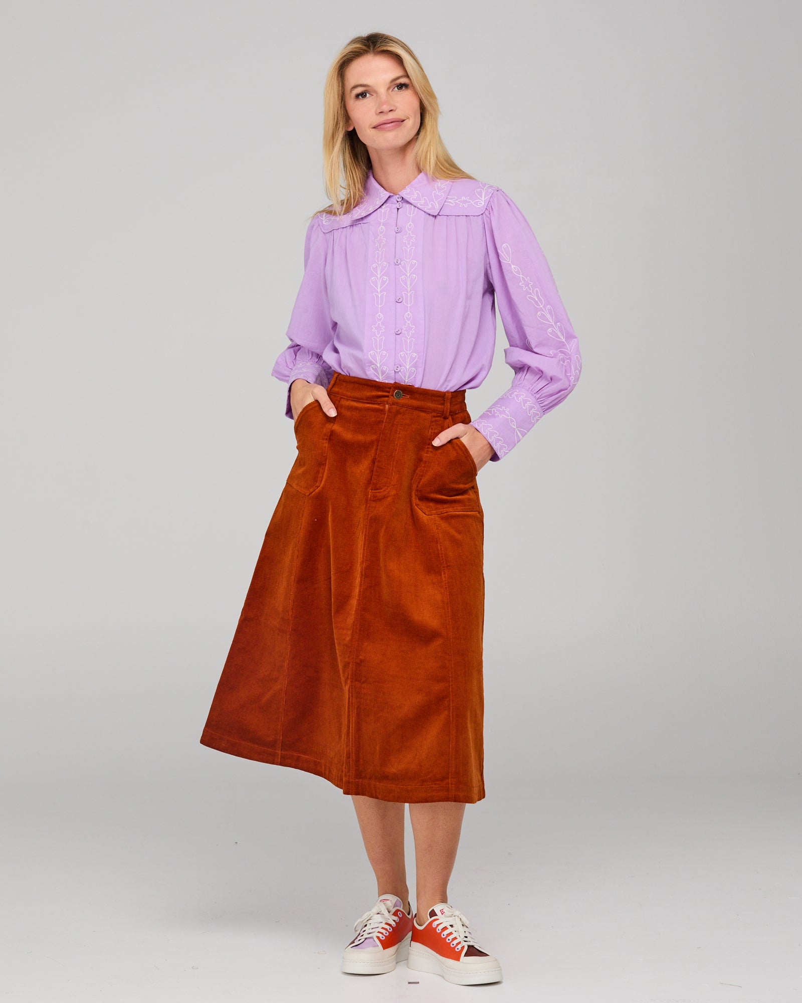 Woman wearing a light purple shirt and rust-colored corduroy skirt on a gray background