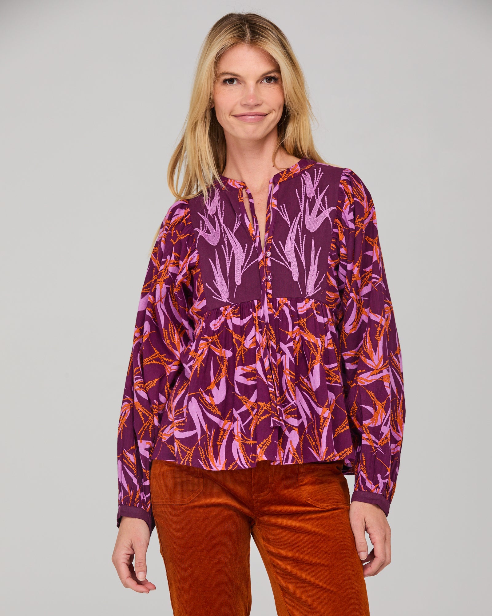 Woman wearing a purple and orange patterned blouse and rust-colored cord pants on a gray background