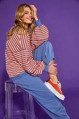 Woman sitting on a stool wearing a red and white striped sweater and blue guru track pants against a purple background