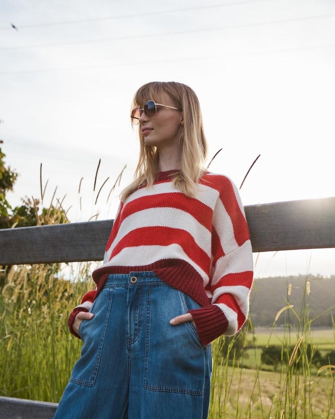 A woman wearing a red and white striped knit sweater and blue denim jeans stands outdoors in a field with a wooden fence behind her.