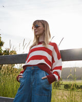 A woman wearing a red and white striped knit sweater and blue denim jeans stands outdoors in a field with a wooden fence behind her.